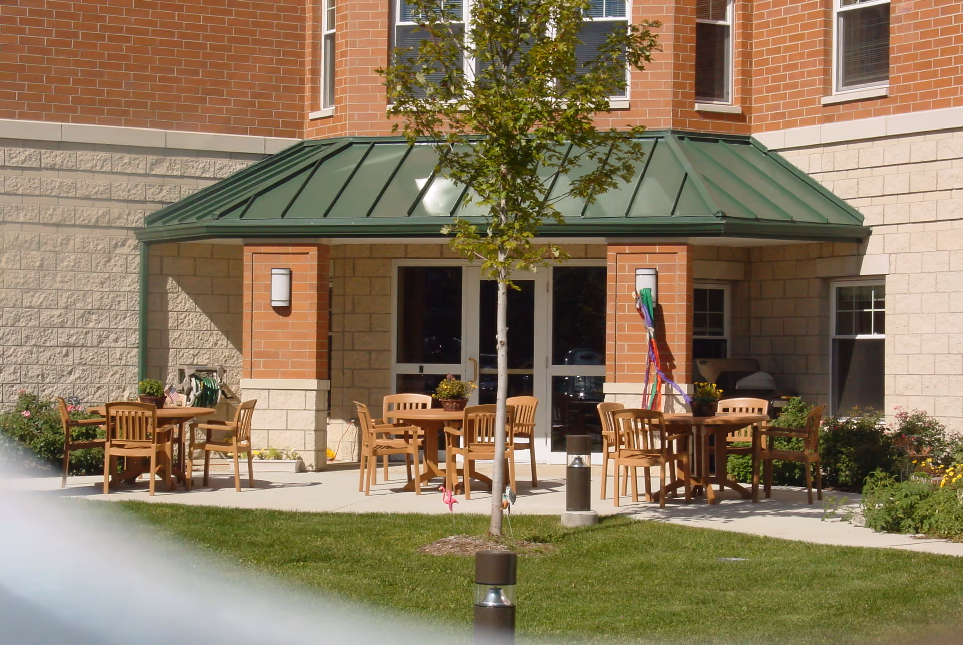 Outdoor patio area at St. Joseph Village of Chicago with wooden tables and chairs arranged on a concrete surface, surrounded by green grass and plants. The building exterior features brick and stone walls with a green metal awning above the entrance doors. A small tree is planted in the grassy area in front of the patio.
