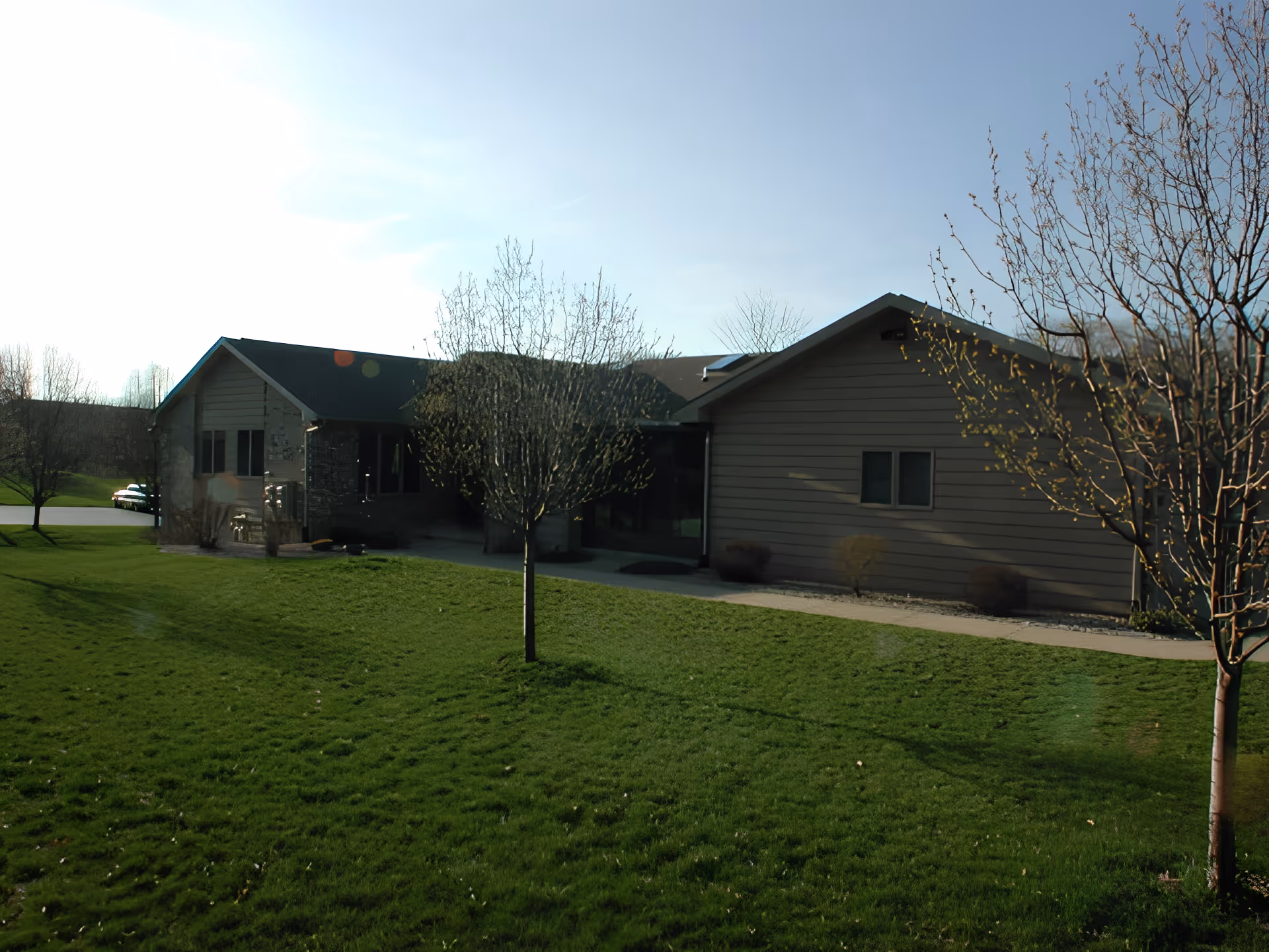 Exterior view of a single-story building with a sloped roof, surrounded by green grass and leafless trees under a clear sky.