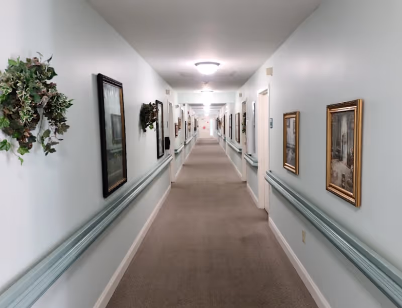 Long carpeted interior hallway with handrails, framed artwork, and ceiling lights in a senior living facility.