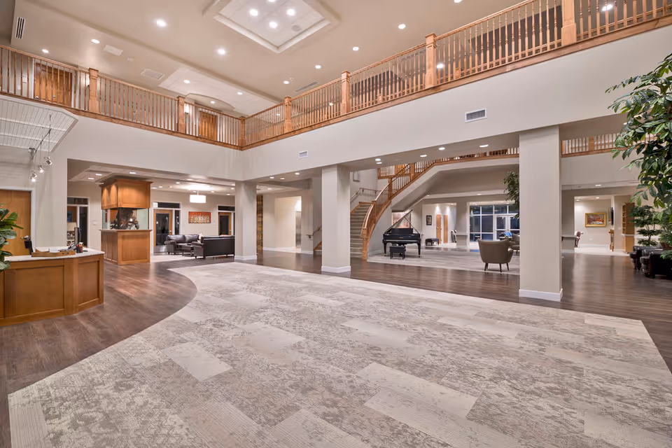 Spacious two-story senior living lobby with a reception desk, grand piano, seating areas and a wooden balcony railing.