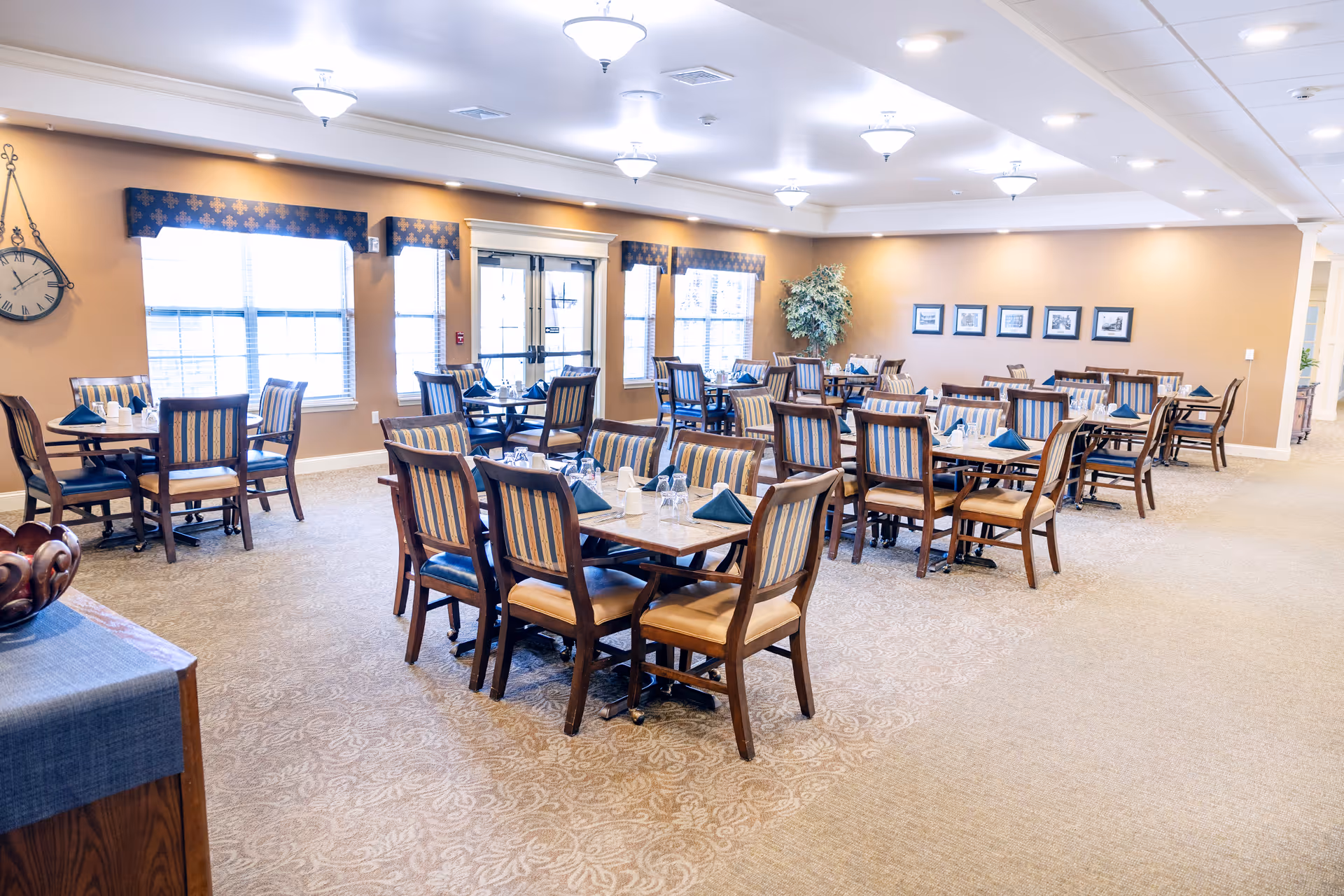 A spacious dining room with multiple wooden tables and chairs arranged neatly. Each table is set with glasses, napkins, and utensils. The room has beige walls, patterned carpet, large windows with blue valances, and ceiling lights providing bright illumination. There are framed pictures on one wall and a large clock on another.