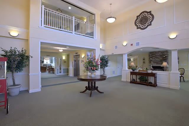 Spacious and well-lit interior common area with high ceilings, a round wooden table with a floral arrangement in the center, potted plants, a decorative wall piece, and a view into adjoining rooms including a dining area and a room with a fireplace.