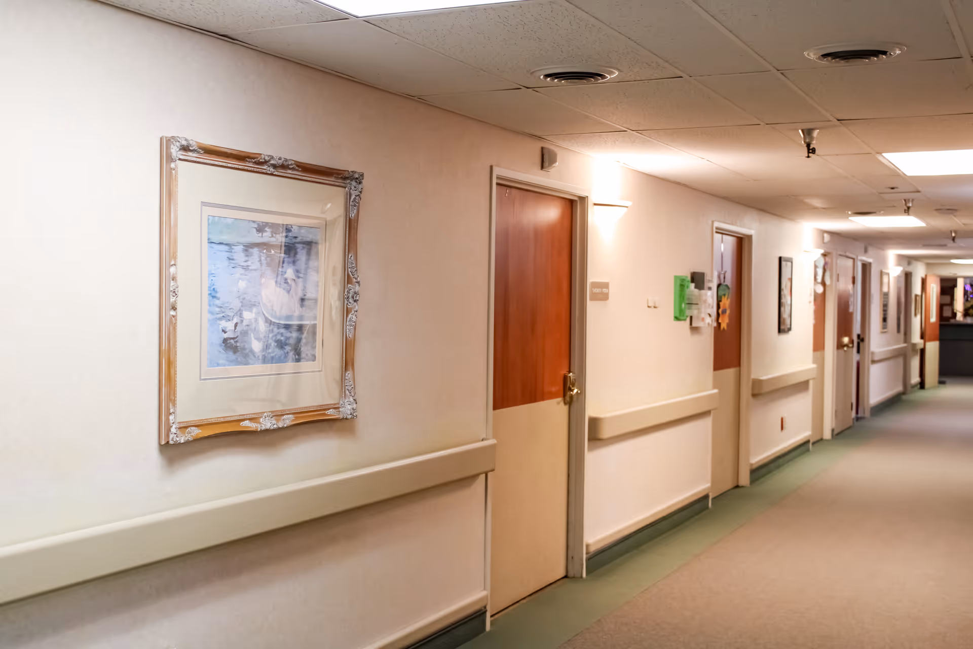 Well-lit interior hallway of a senior living facility with closed wooden doors, handrails, and framed artwork on the walls.