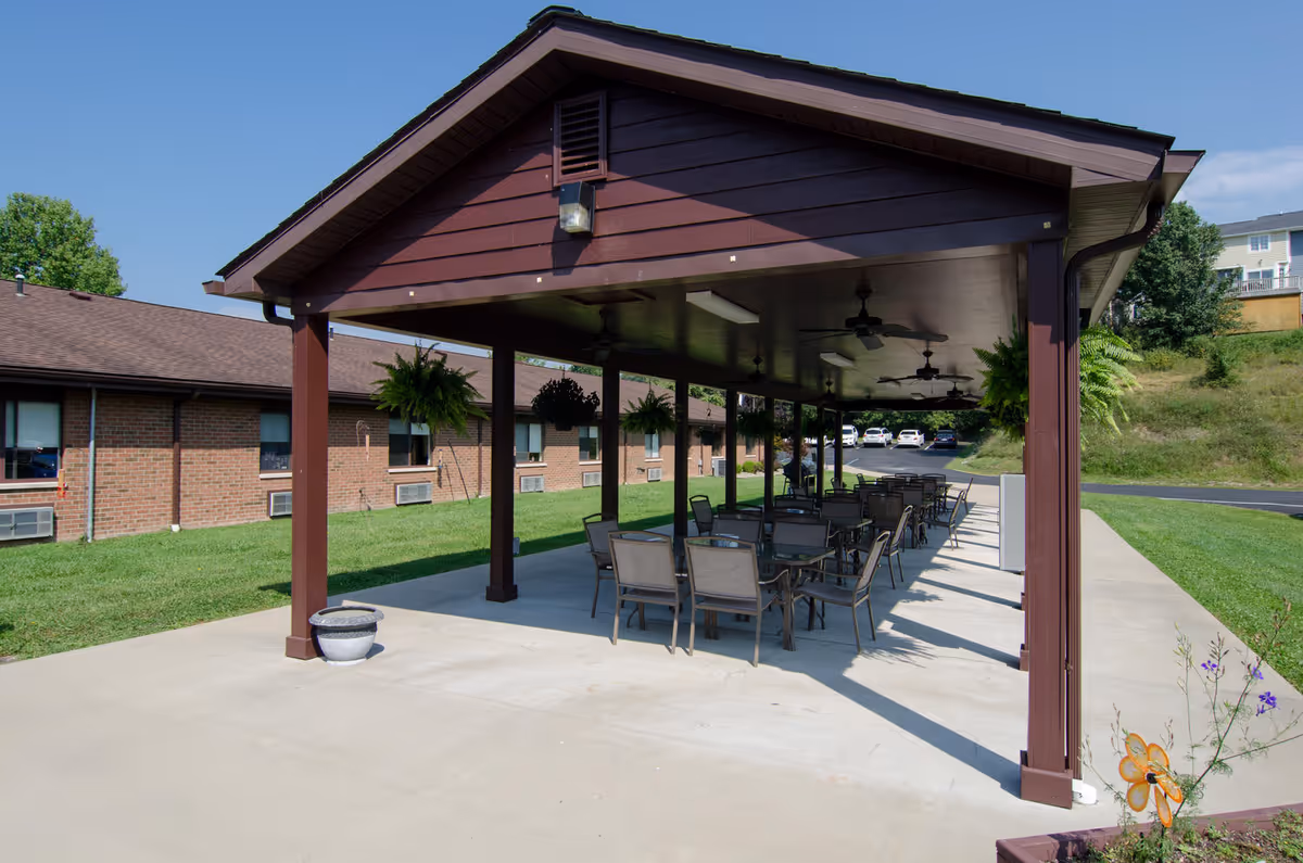 Outdoor covered patio area with multiple tables and chairs arranged underneath a brown wooden pavilion. Hanging plants are suspended from the ceiling, and ceiling fans are installed for ventilation. The patio is adjacent to a brick building with windows, and there is a grassy area surrounding the patio. Several cars are parked in the background on a paved driveway.