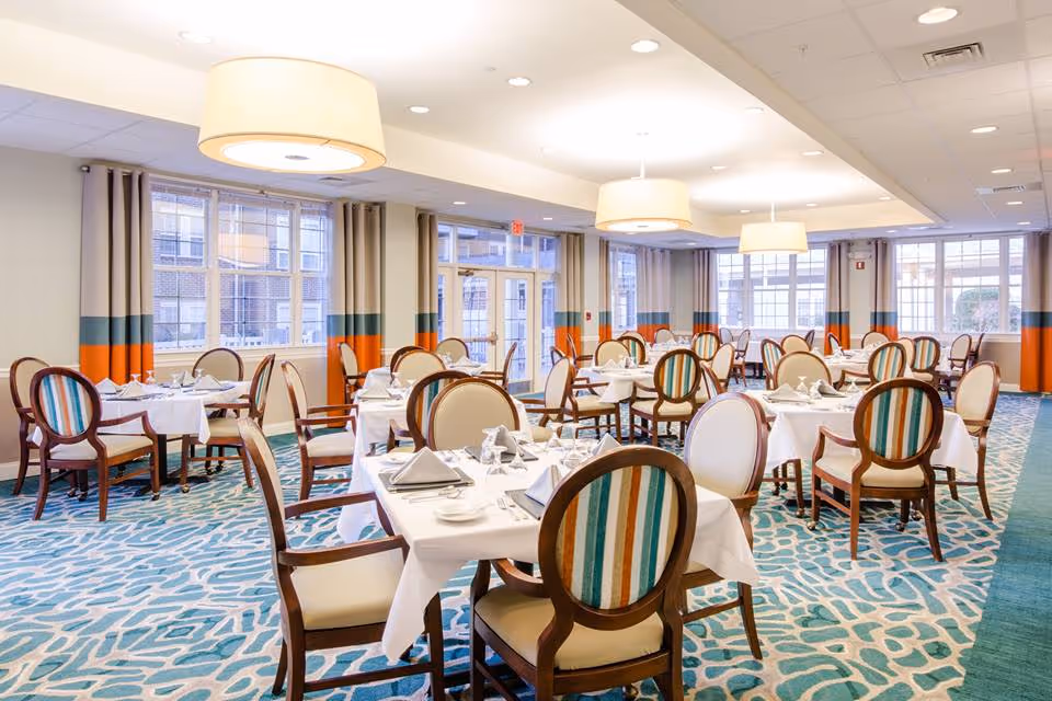 A bright and spacious dining room with multiple round tables covered in white tablecloths, each set with napkins, plates, and glassware. The chairs have wooden frames with beige and striped upholstery. Large windows with curtains allow natural light to fill the room, and modern pendant lights hang from the ceiling.