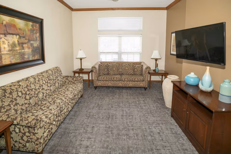 A living room with two patterned sofas facing each other, a large window with blinds in the center, two side tables each with a lamp, a wooden TV stand with decorative vases, and a wall-mounted flat-screen TV. A framed painting hangs on the left wall.