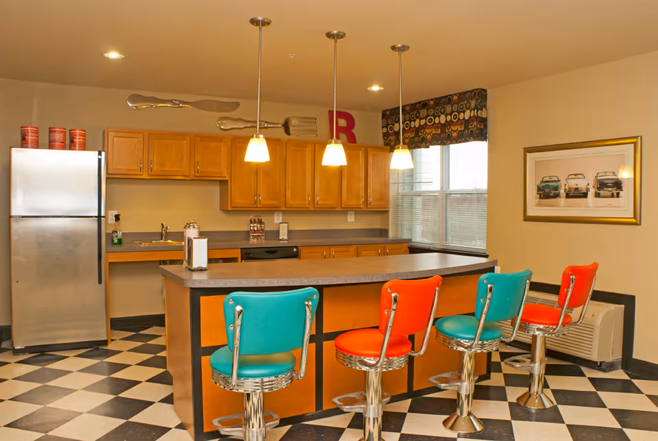 A kitchen area with a checkered black and white floor, a large island with a gray countertop, and four retro-style bar stools in alternating teal and red colors. The kitchen has wooden cabinets, a stainless steel refrigerator, a sink, and three pendant lights hanging above the island. A window with a patterned valance is on the right wall, which also features a framed picture of vintage cars.