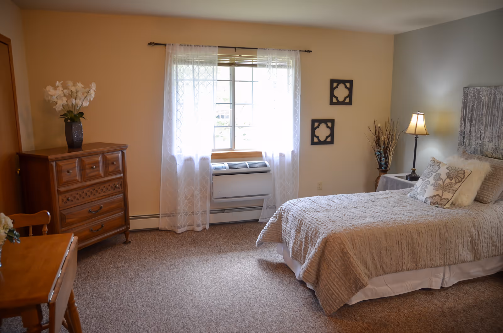 A bright, neatly decorated single bedroom with a bed, nightstand, dresser and a window with sheer curtains.