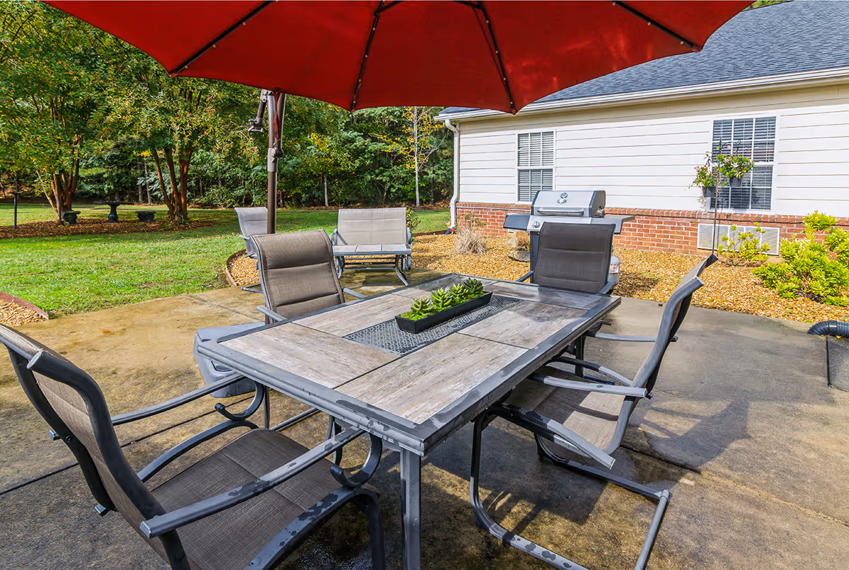 Outdoor patio area with a rectangular table surrounded by six chairs under a large red umbrella. There is a small rectangular planter with green succulents on the table. In the background, there is a white building with brick trim, a barbecue grill, and some greenery including trees and bushes.