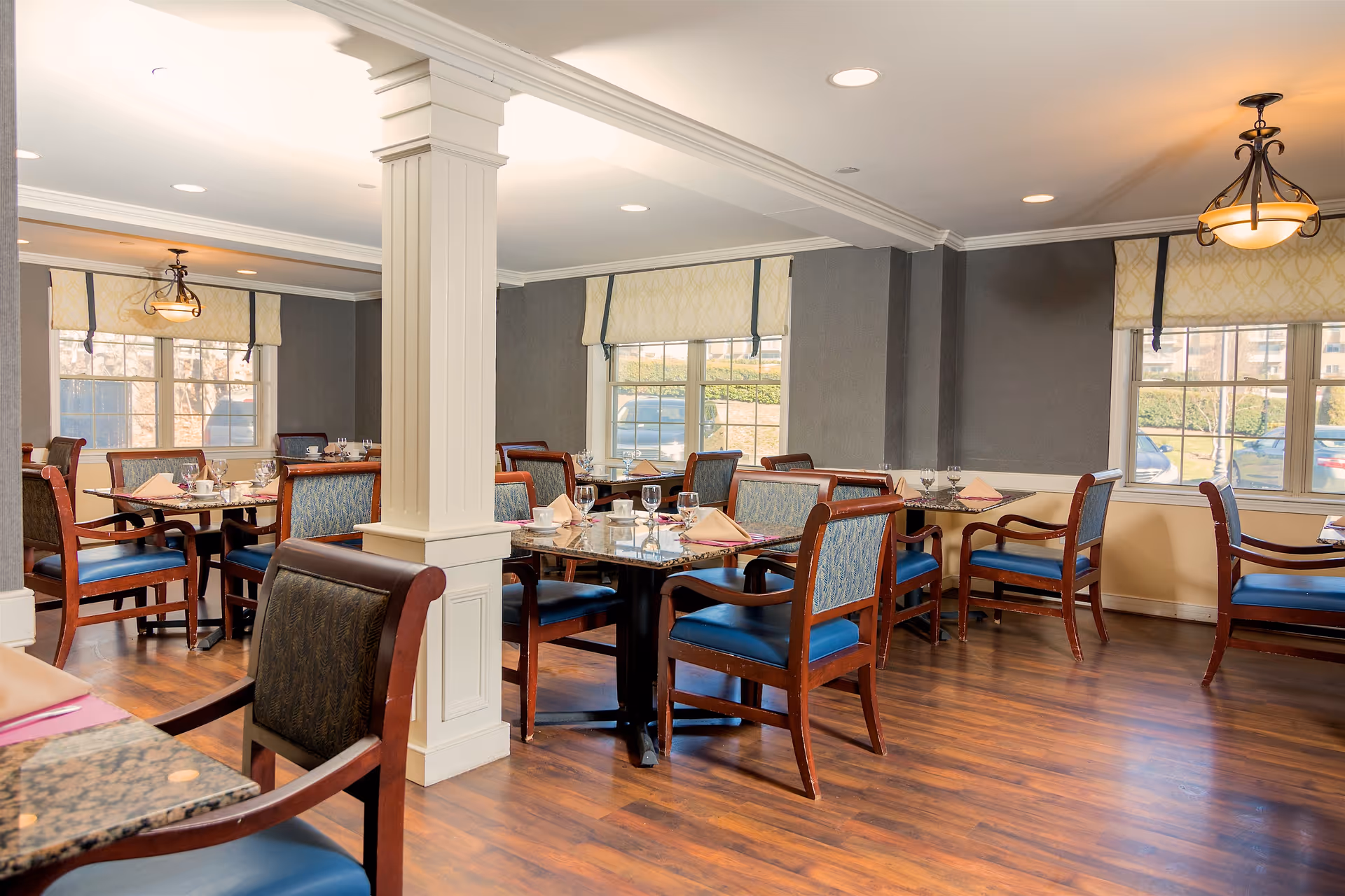 Bright dining room with multiple set tables and wooden chairs around a central white column and large windows.