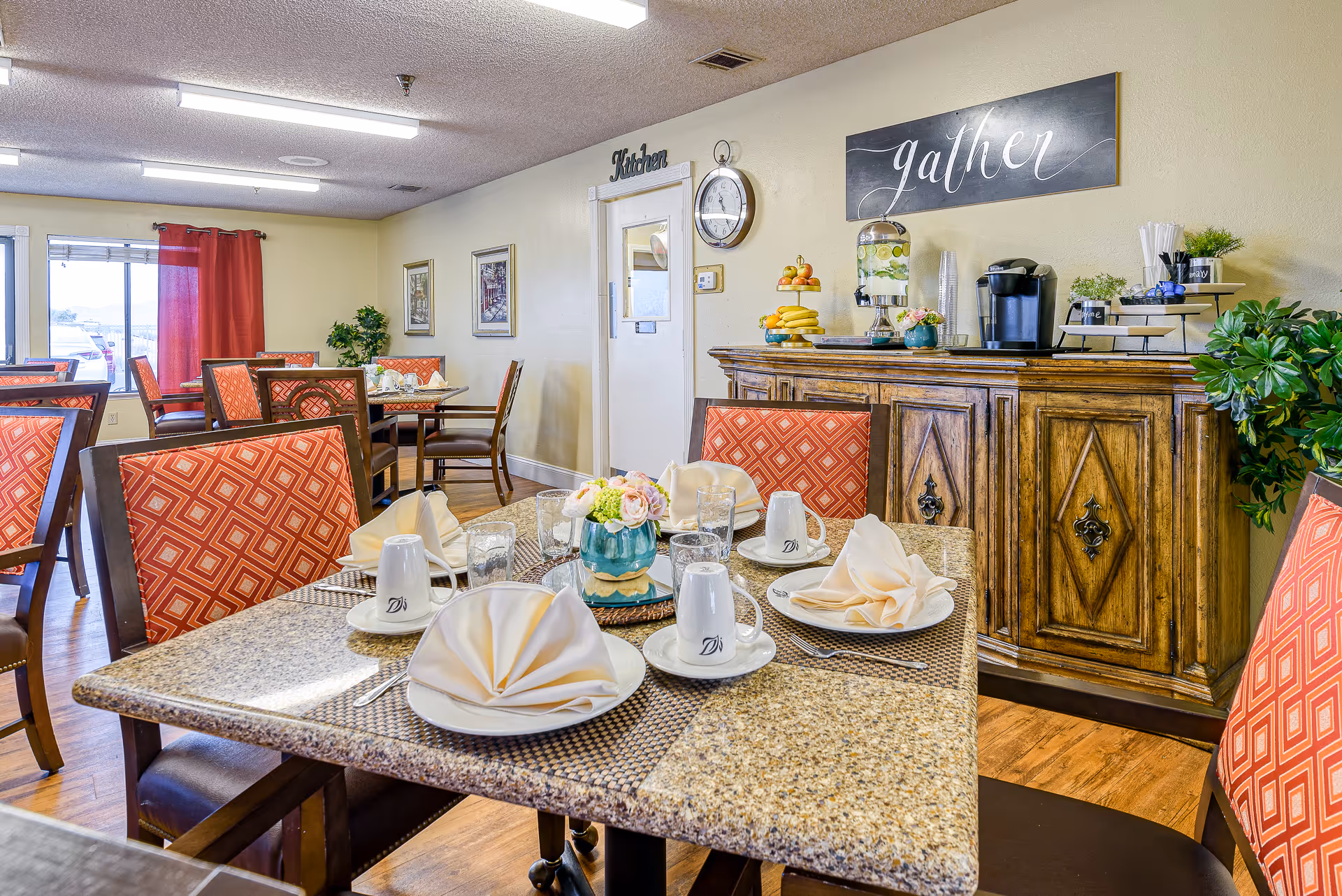 Dining area with tables set for a meal, featuring granite tabletops, folded napkins on plates, cups, and a small flower arrangement. The room has red patterned chairs, wooden flooring, and a sideboard with a coffee maker, fruit, and a water dispenser. A sign on the wall reads 'gather' and another above a door reads 'Kitchen'.