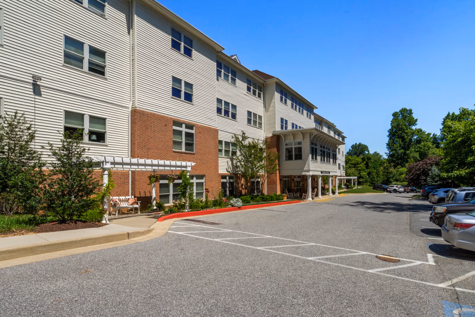 Front exterior of a multi-story senior living facility showing the entrance, parking lot, and surrounding trees under a clear blue sky.