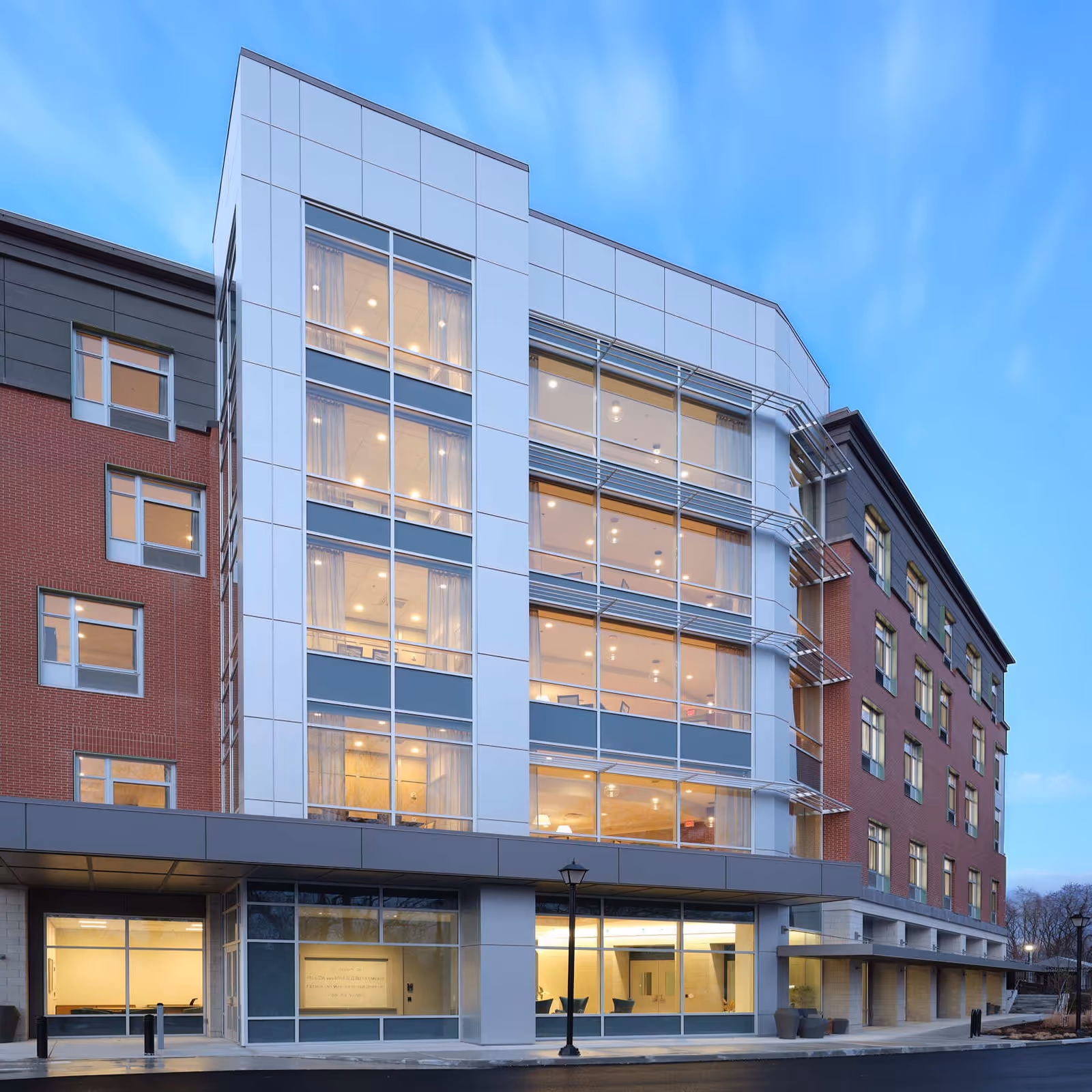 Exterior view of a modern multi-story senior living facility building with large windows and a combination of brick and metal paneling under a clear blue sky.