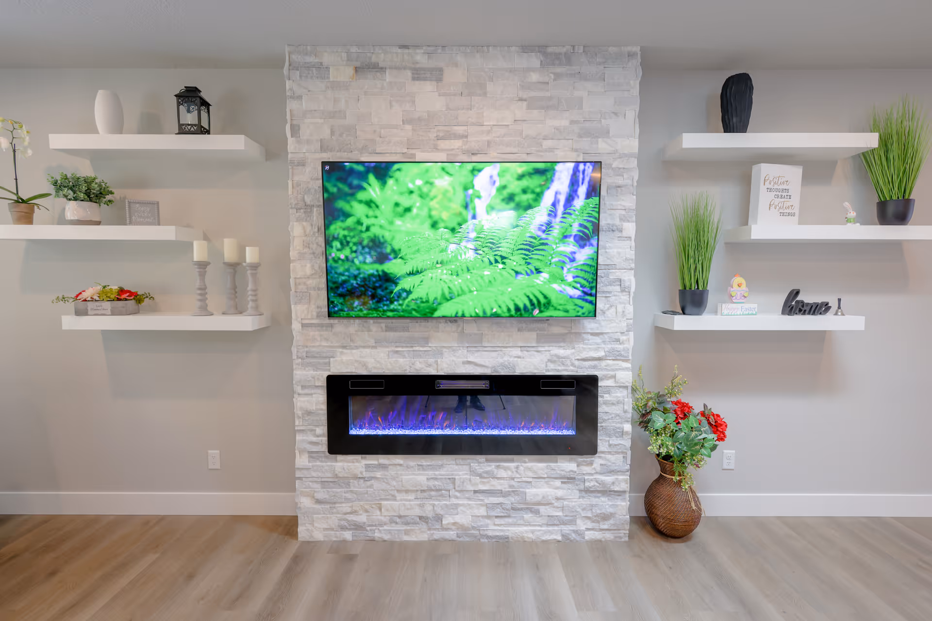 Modern living room wall with a mounted TV above a stone electric fireplace flanked by white floating shelves holding plants and decor.