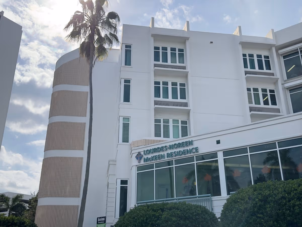 White multi-story senior residence building entrance with large windows, landscaping, a tall palm tree, and a sign for the McKeen Residence.