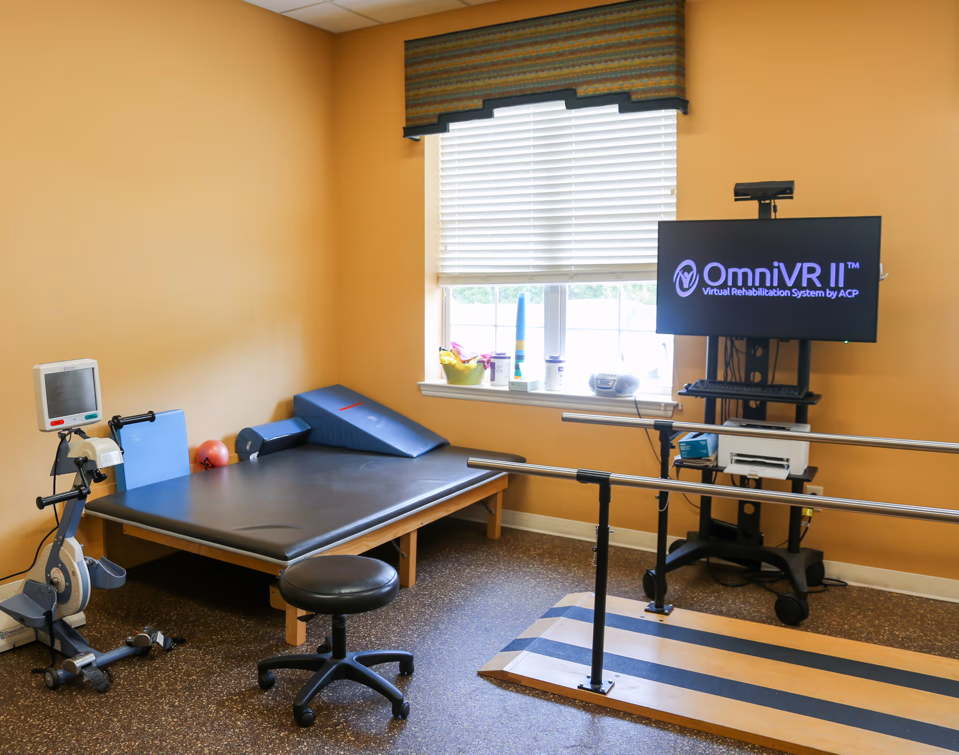 A rehabilitation therapy room with a padded therapy table, exercise bike, parallel bars for walking practice, a stool, and a monitor displaying 'OmniVR II Virtual Rehabilitation System by ACP'. The room has yellow walls, a window with blinds and a valance, and various therapy equipment on the window sill.