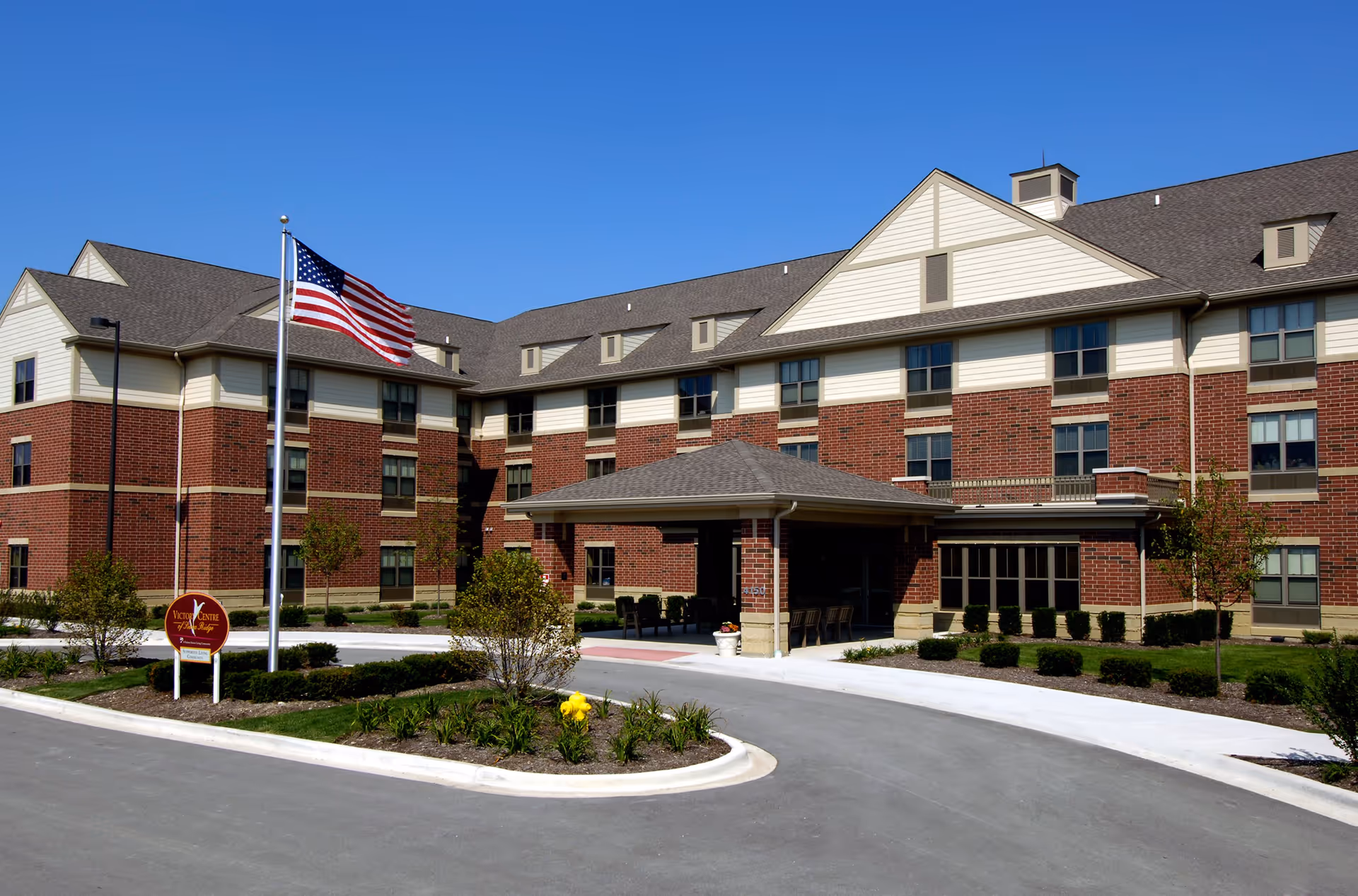 Exterior view of Victory Centre of Sierra Ridge, a multi-story brick and beige siding building with a covered entrance, an American flag on a flagpole, and landscaped greenery under a clear blue sky.