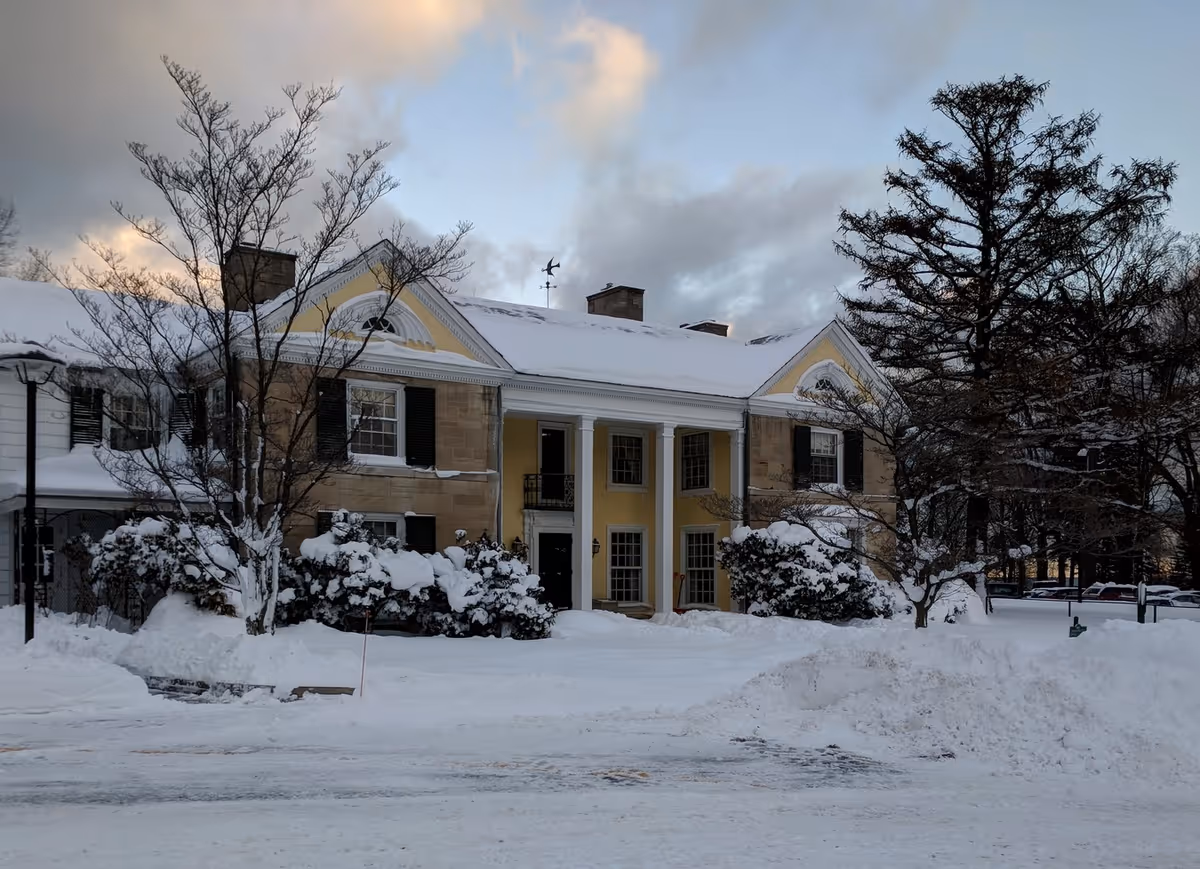 Front view of a two-story classical building with columns and snow-covered grounds under a cloudy sky.