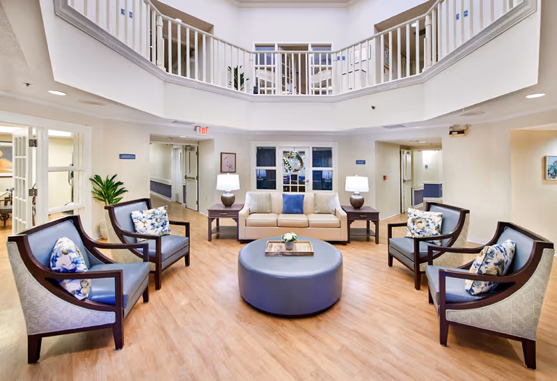 A bright and spacious senior living facility common area with a beige sofa and four armchairs arranged around a large round ottoman. The chairs have floral patterned cushions. Two side tables with lamps flank the sofa. The room has light wood flooring and white walls with a balcony railing visible on the upper level. There are plants and framed artwork on the walls.