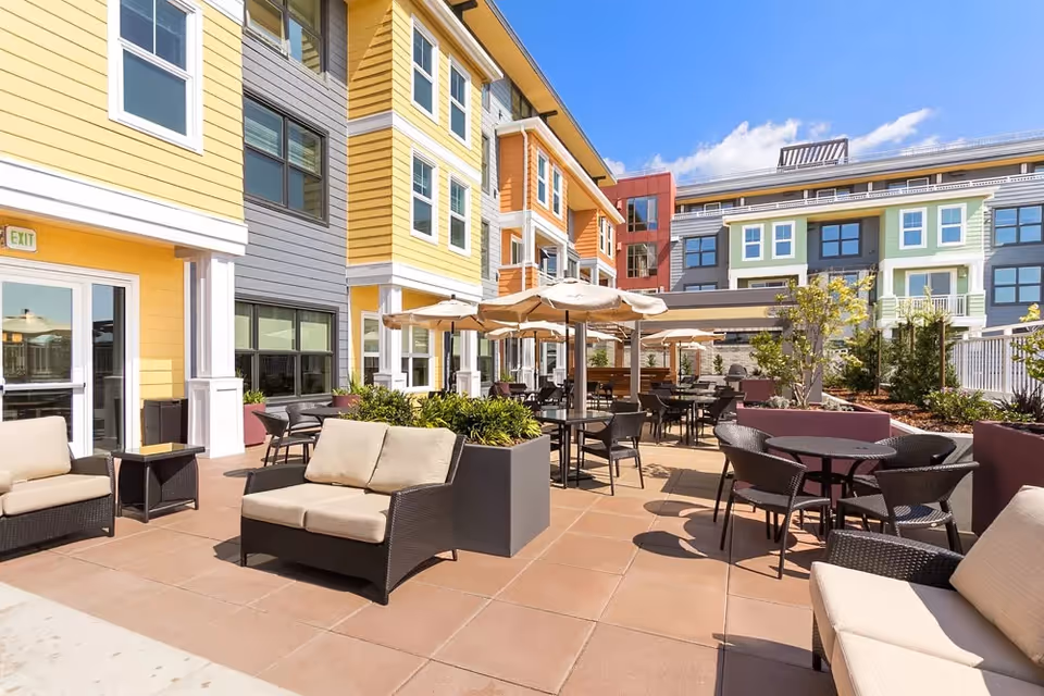 Outdoor patio area at Merrill Gardens at Rockridge featuring cushioned wicker seating, round tables with umbrellas, potted plants, and a multi-story building with colorful siding in the background under a clear blue sky.