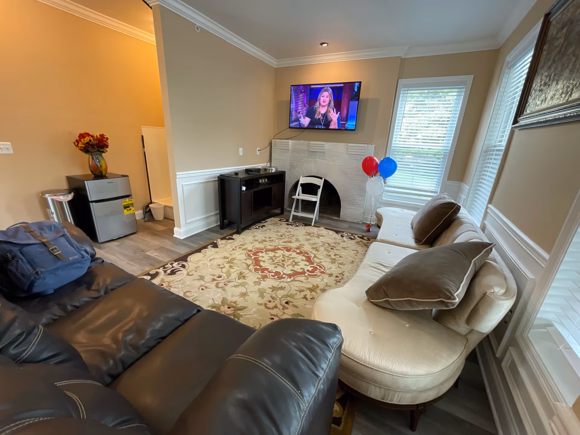 A cozy living room with a black leather sofa and a cream-colored curved sofa with brown pillows. A patterned area rug covers the floor. A flat-screen TV is mounted above a white brick fireplace. There is a small black cabinet next to the fireplace and a cluster of red, white, and blue balloons near the window. A small refrigerator and a vase with flowers are visible in an adjacent area.