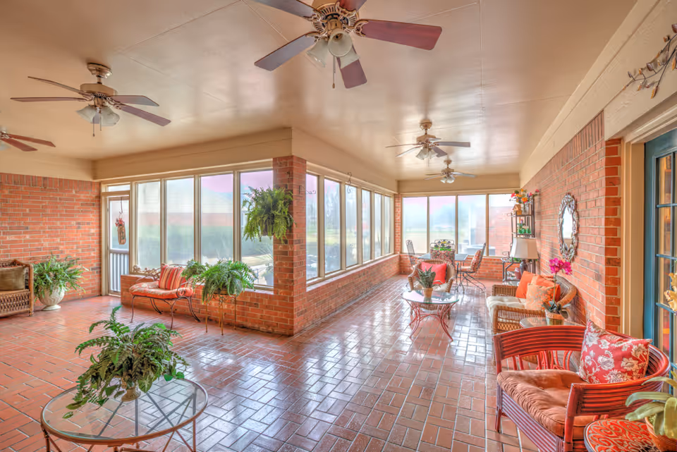 A bright and spacious sunroom with large windows along two walls, allowing natural light to fill the space. The room features brick walls and a tiled floor, with several ceiling fans overhead. There are multiple seating areas with cushioned chairs, wicker furniture, and small tables adorned with potted plants and decorative items. The room has a cozy and inviting atmosphere with various plants placed throughout.