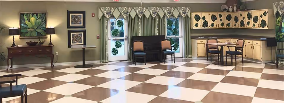 Spacious interior common room with a brown-and-white checkered floor, tables and chairs, cabinets, piano, and wall artwork.