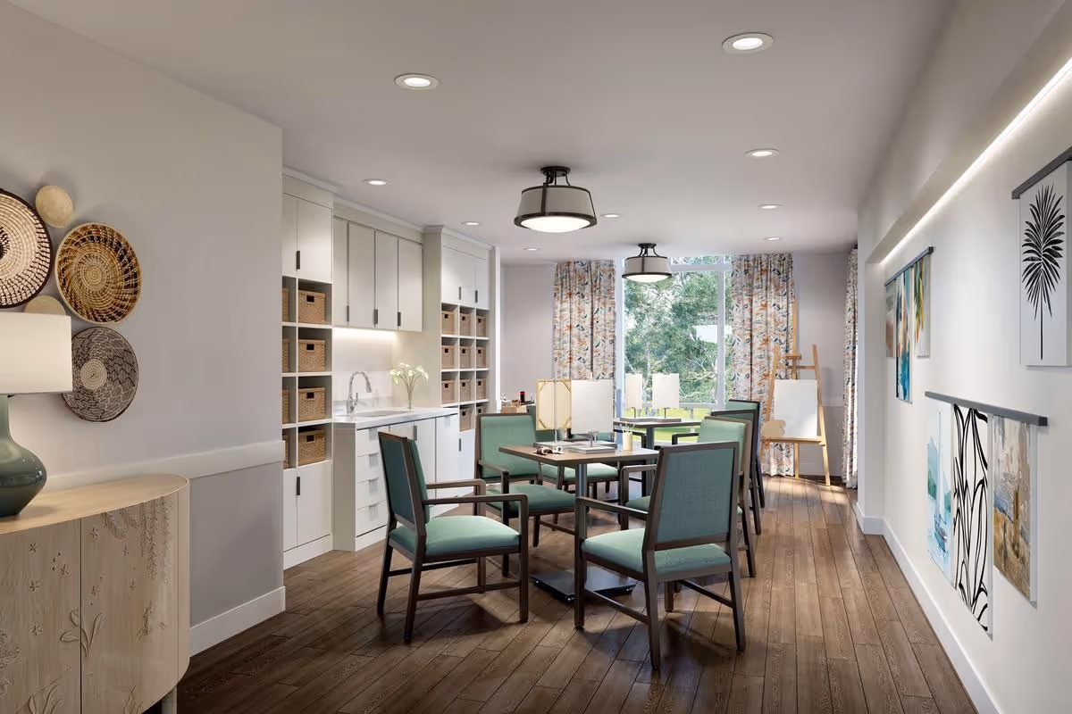 Well-lit common dining/activity room with a central table surrounded by teal-upholstered chairs, a kitchenette with storage baskets, and wall artwork.