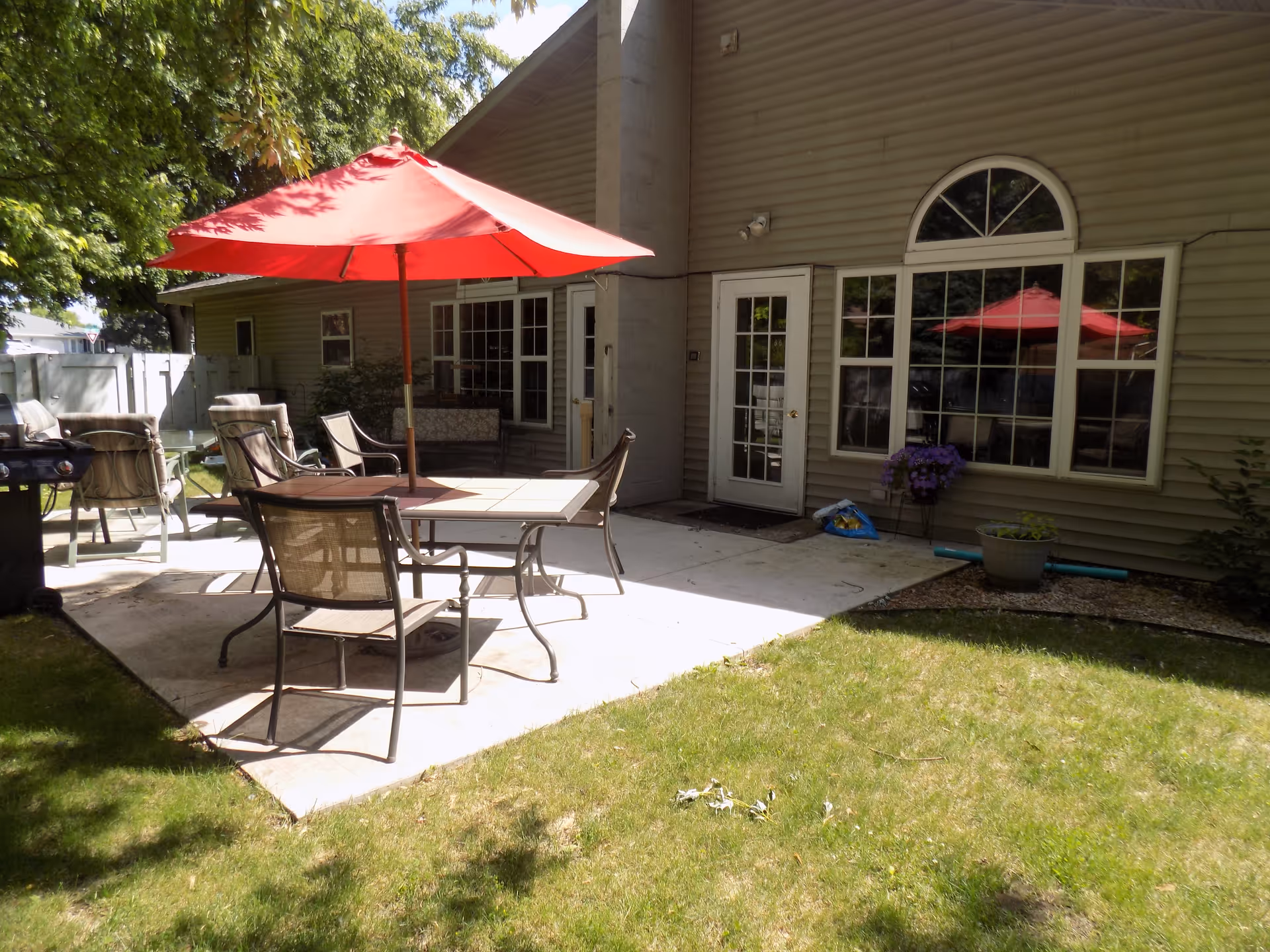Outdoor patio area with a table and four chairs under a red umbrella, surrounded by green grass and trees. The patio is adjacent to a building with beige siding, large windows, and a door. There are additional chairs and a grill on the patio.