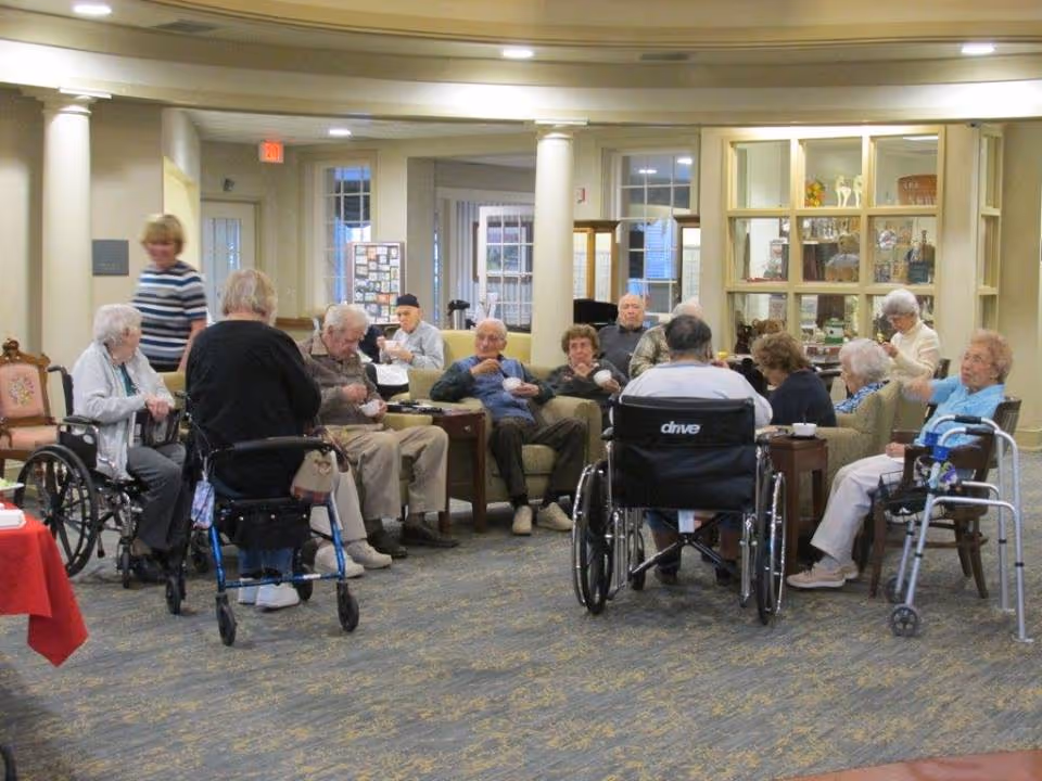 A group of elderly people sitting and socializing in a common area of a senior living facility. Some are seated in wheelchairs, others in chairs and on sofas. A staff member is standing nearby. The room has columns, carpeted floor, and a display cabinet in the background.