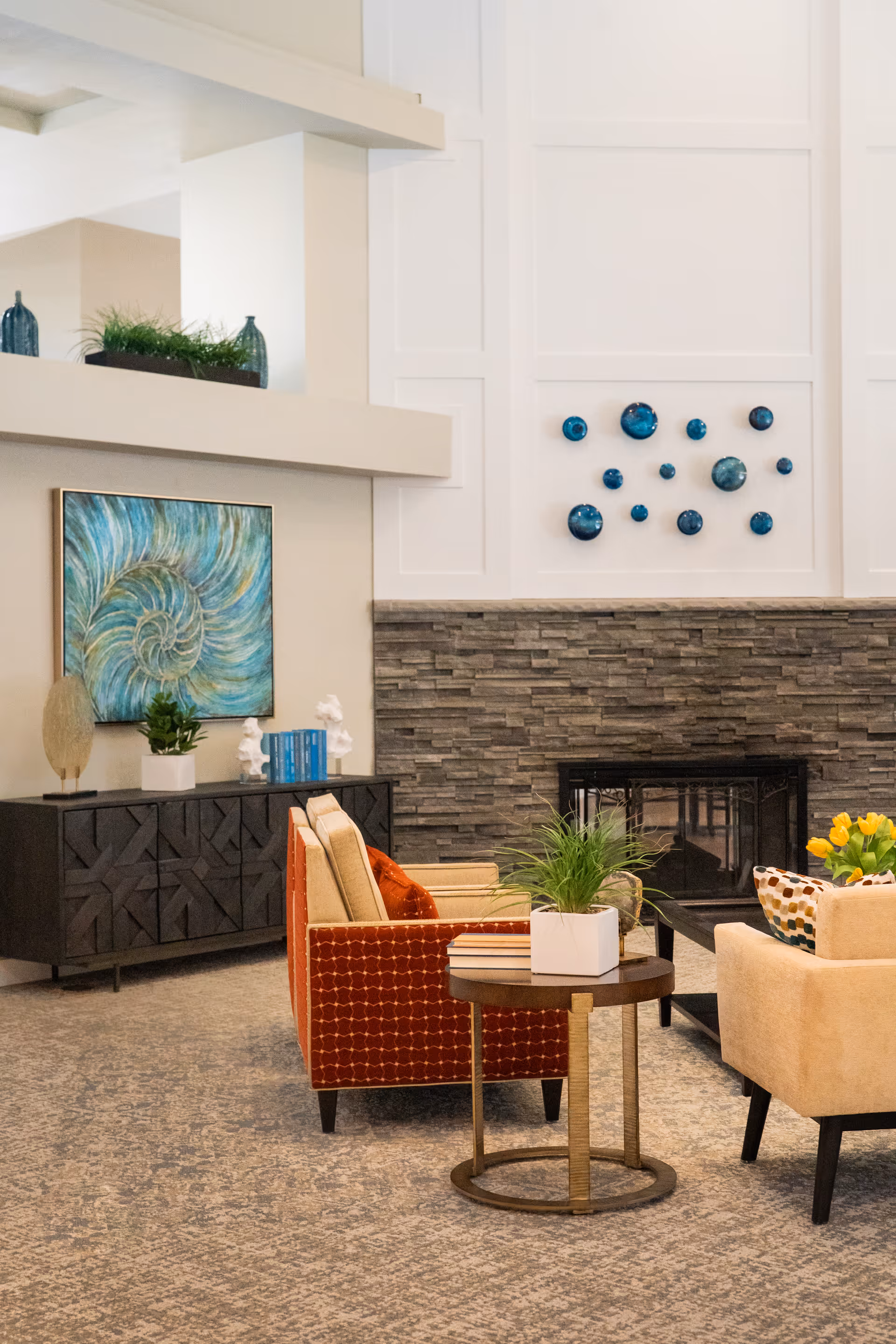 A cozy living room area with a stone fireplace, decorated with blue circular wall art above it. The room features a patterned orange armchair, a beige armchair with a colorful pillow, and a round wooden side table with a potted plant and books. A dark wooden cabinet with decorative items and a blue abstract painting is against the wall.