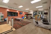A bright therapy/fitness room with exercise machines, a treatment table, colorful exercise balls, and a wall-mounted TV beneath a 'Strength Through Recovery' sign.