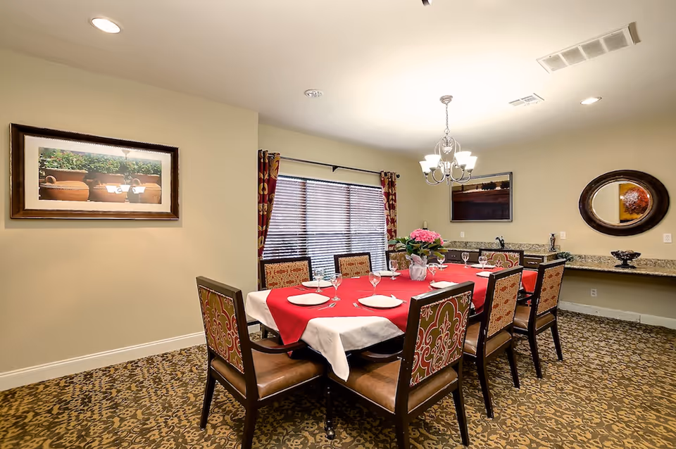 A dining room with a rectangular table set for eight with a red tablecloth, patterned chairs, chandelier, and wall decor.
