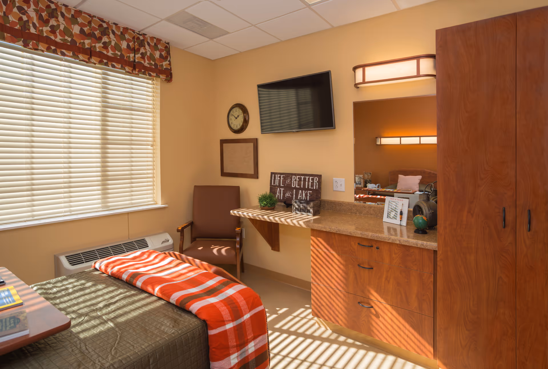 Sunlit senior living bedroom with a bed, chair, wall-mounted TV, countertop dresser and wardrobe.