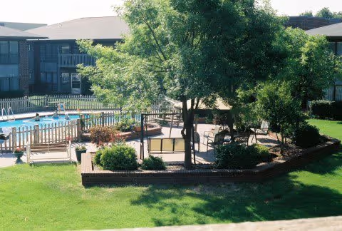 Outdoor area of a senior living facility featuring a swimming pool surrounded by a white fence, a patio with chairs and tables under a large tree, and well-maintained green grass and shrubs. The background shows a multi-story building with balconies.