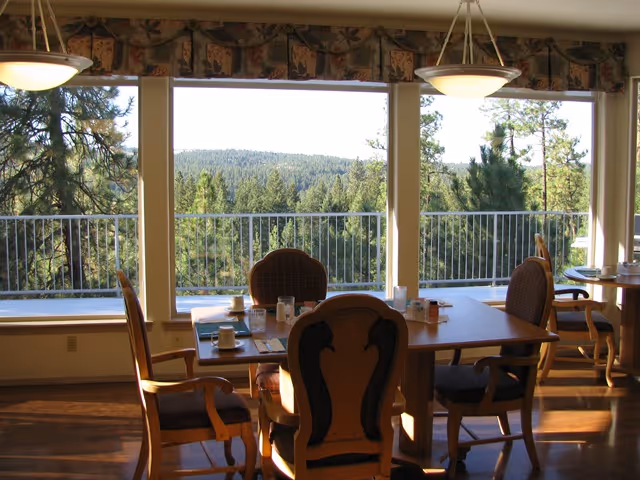 Dining area with wooden tables and chairs set with cups and glasses, large windows offering a view of a forested landscape and hills in the distance.
