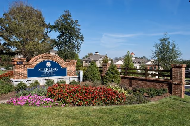 Entrance sign for Sterling Estates Senior Living Community surrounded by landscaped flowers, shrubs, and a green lawn with a clear blue sky in the background.