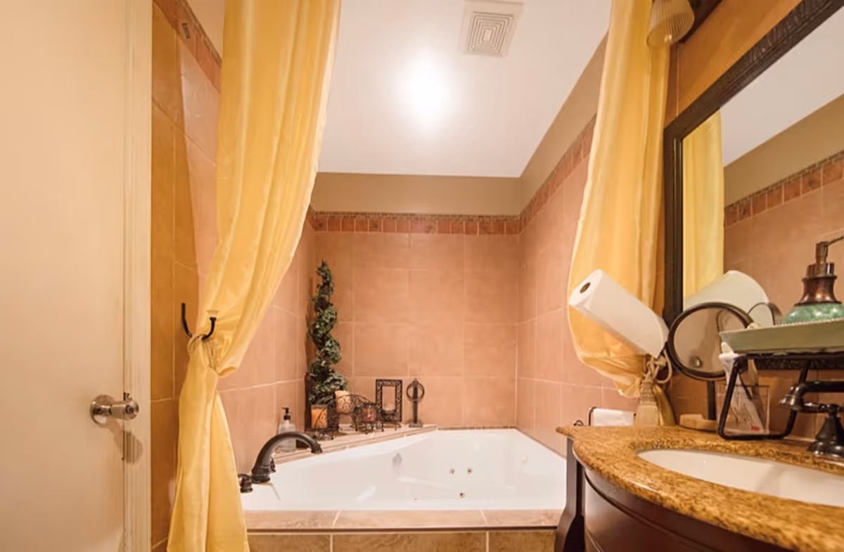 A bathroom with a large white bathtub surrounded by tan tiles and a decorative border. Yellow curtains are tied back on either side of the tub. To the right, there is a granite countertop with a sink, a mirror, and various toiletries including a paper towel roll and soap dispenser. A small green plant decoration is placed on the ledge behind the tub.