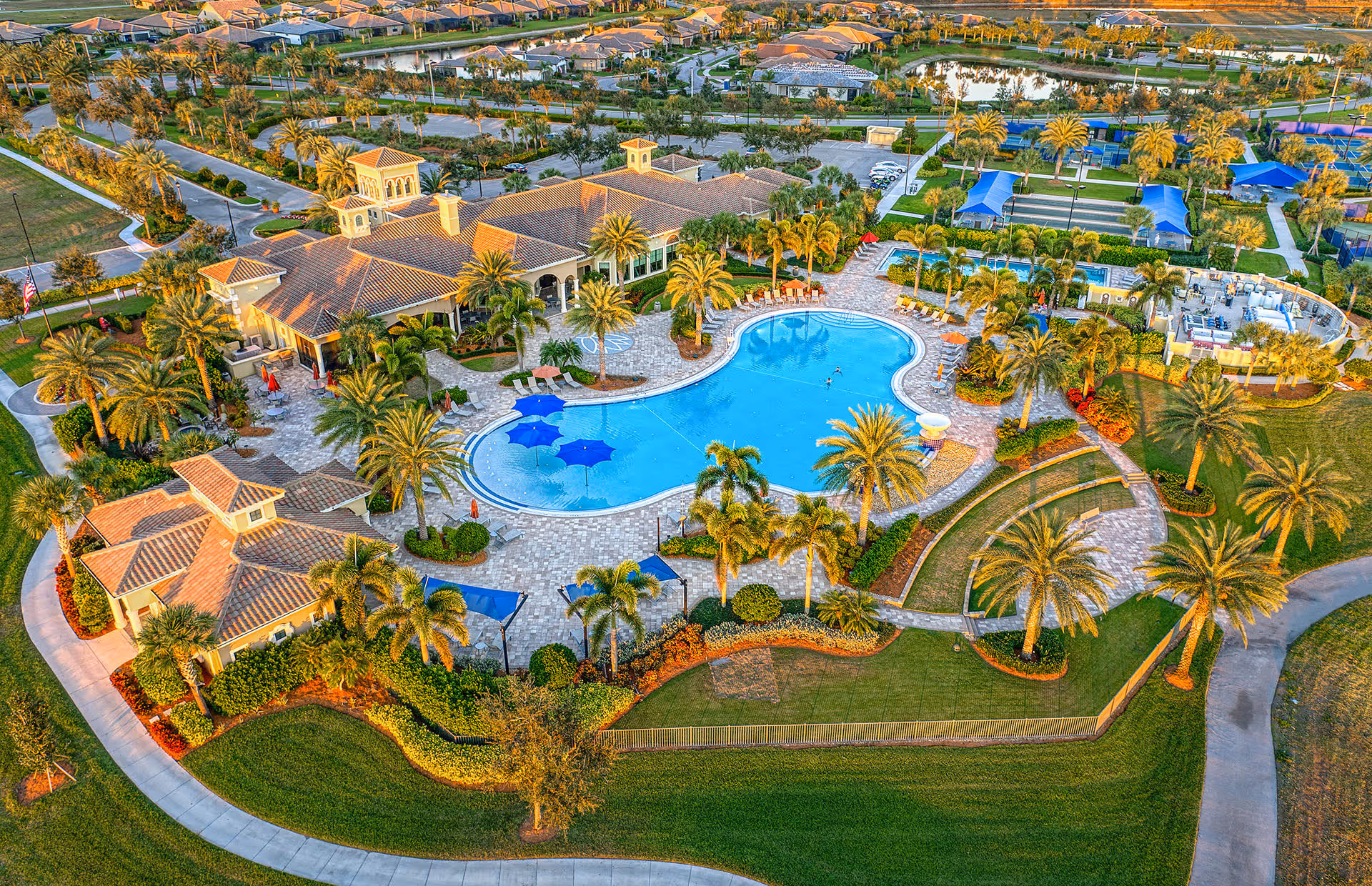 Aerial view of Del Webb Naples 55+ Retirement Community showing a large swimming pool surrounded by palm trees, lounge chairs, and umbrellas. The pool area is adjacent to a clubhouse with a tiled roof and multiple buildings. There are landscaped gardens, walking paths, and recreational areas including tennis courts with blue canopies. Residential houses and a pond are visible in the background.