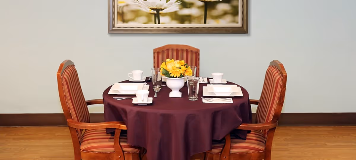 A round dining table covered with a dark purple tablecloth set for four people with white square plates, cups, glasses, and silverware. A white vase with yellow flowers is placed in the center of the table. Four wooden chairs with red and gold striped upholstery surround the table. A framed picture of white flowers hangs on the light-colored wall behind the table.