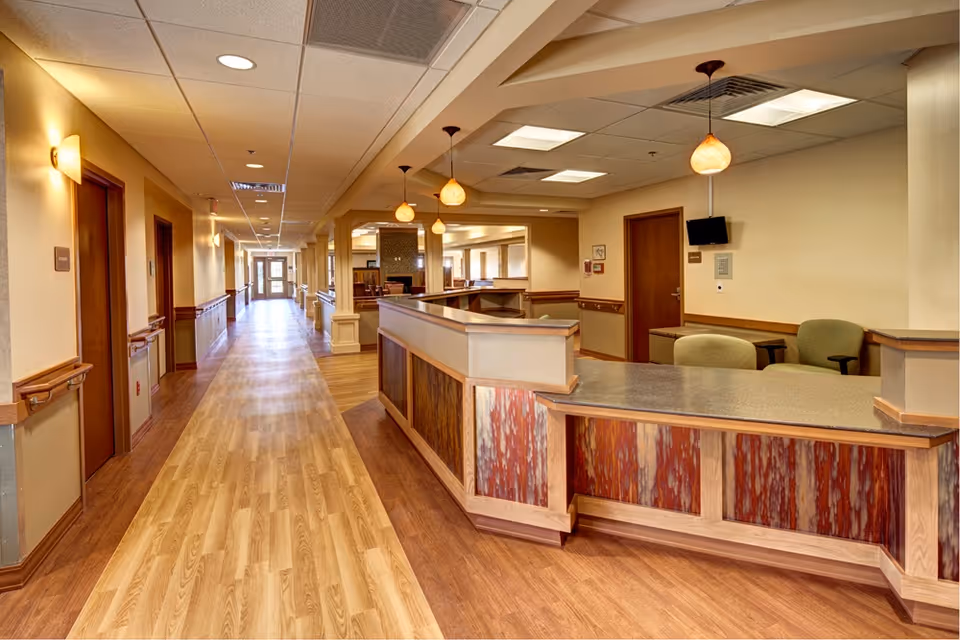 A wide hallway in an assisted living facility with wood flooring and beige walls. On the right side, there is a reception desk with a granite countertop and decorative wood paneling. Behind the desk are two green chairs and a small wall-mounted TV. The hallway has several doors along the left side and is well-lit with ceiling lights and hanging pendant lamps above the reception area.