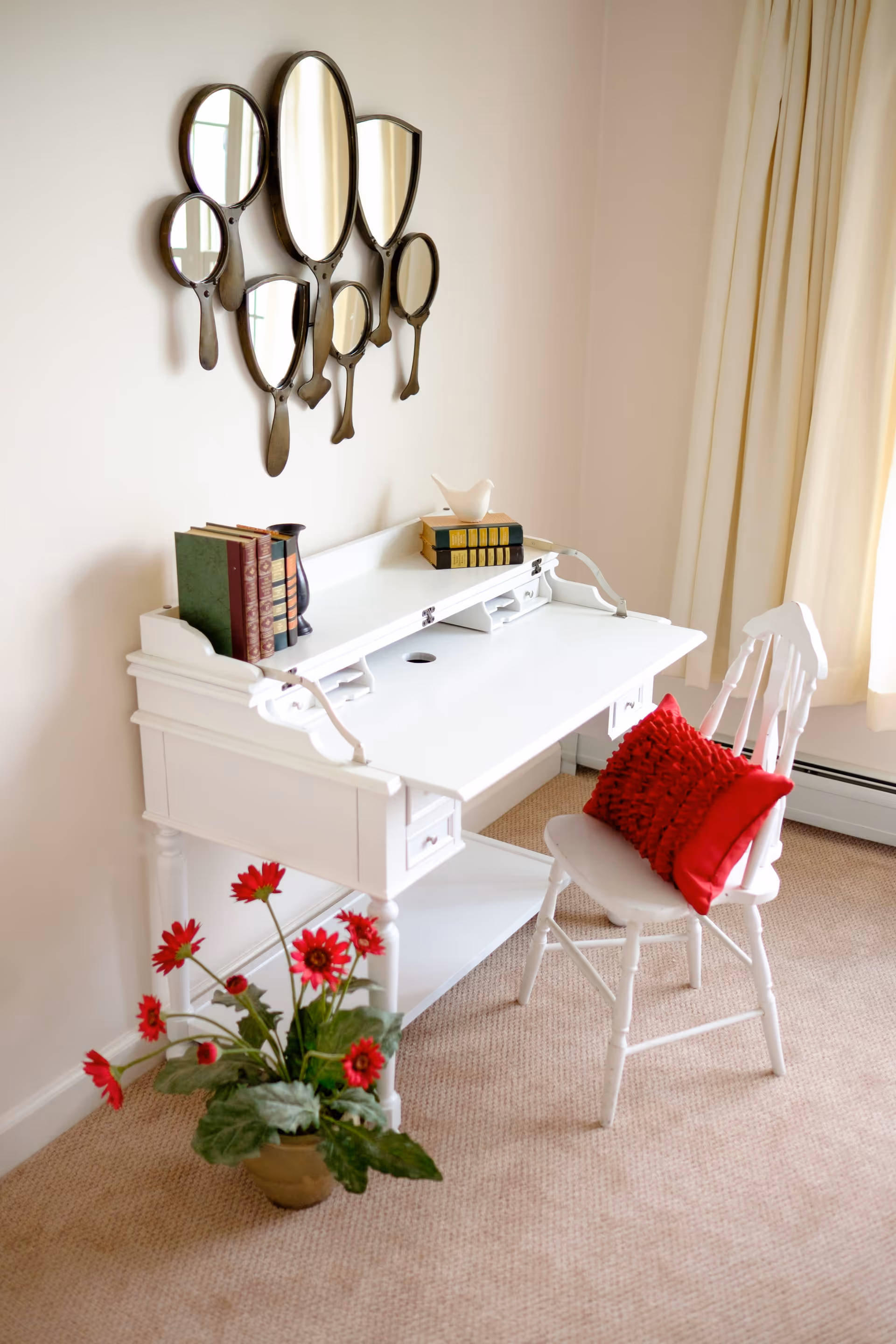 A white wooden desk with small drawers and a white chair with a red cushion in front of it. On the desk are several books, a small black vase, and a white bird figurine. Above the desk is a decorative wall mirror composed of multiple hand mirror shapes. A pot with red flowers is placed on the floor next to the desk. The room has beige carpet and cream-colored curtains.