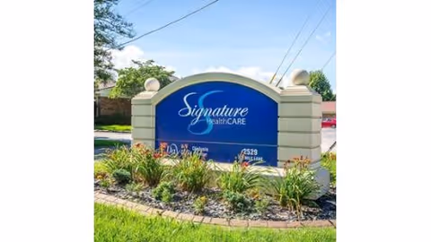 A blue entrance sign reading 'Signature HealthCARE' set between stone pillars with landscaping and a road behind it.
