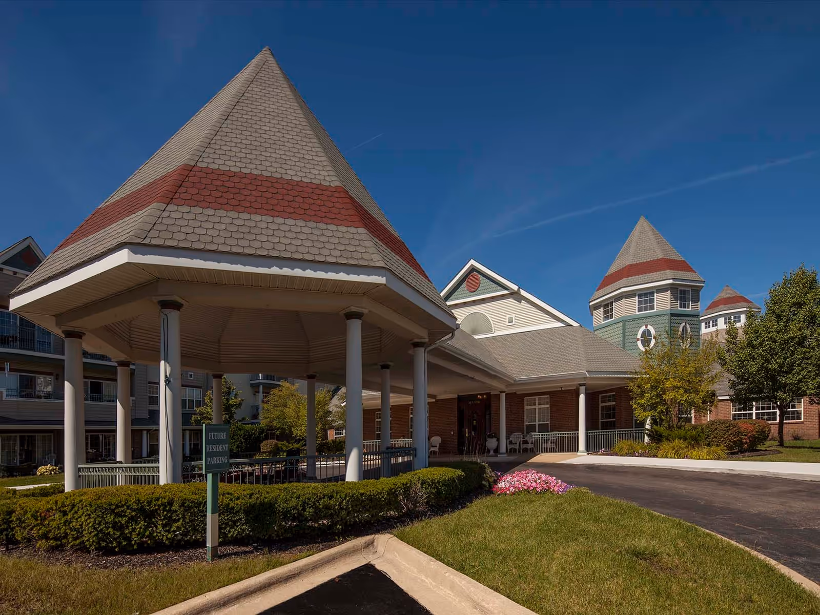 Exterior view of Waltonwood Cherry Hill senior living facility featuring a covered entrance with a conical roof supported by columns, landscaped bushes, a driveway, and a building with multiple peaked roofs and windows under a clear blue sky.