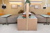 A hospital or nursing facility room with two beds separated by a partial divider. Each bed has a pillow and a small overbed table. Mounted monitors and medical equipment are visible on the walls above the beds. A small cabinet with a flower arrangement is positioned in the center between the beds.