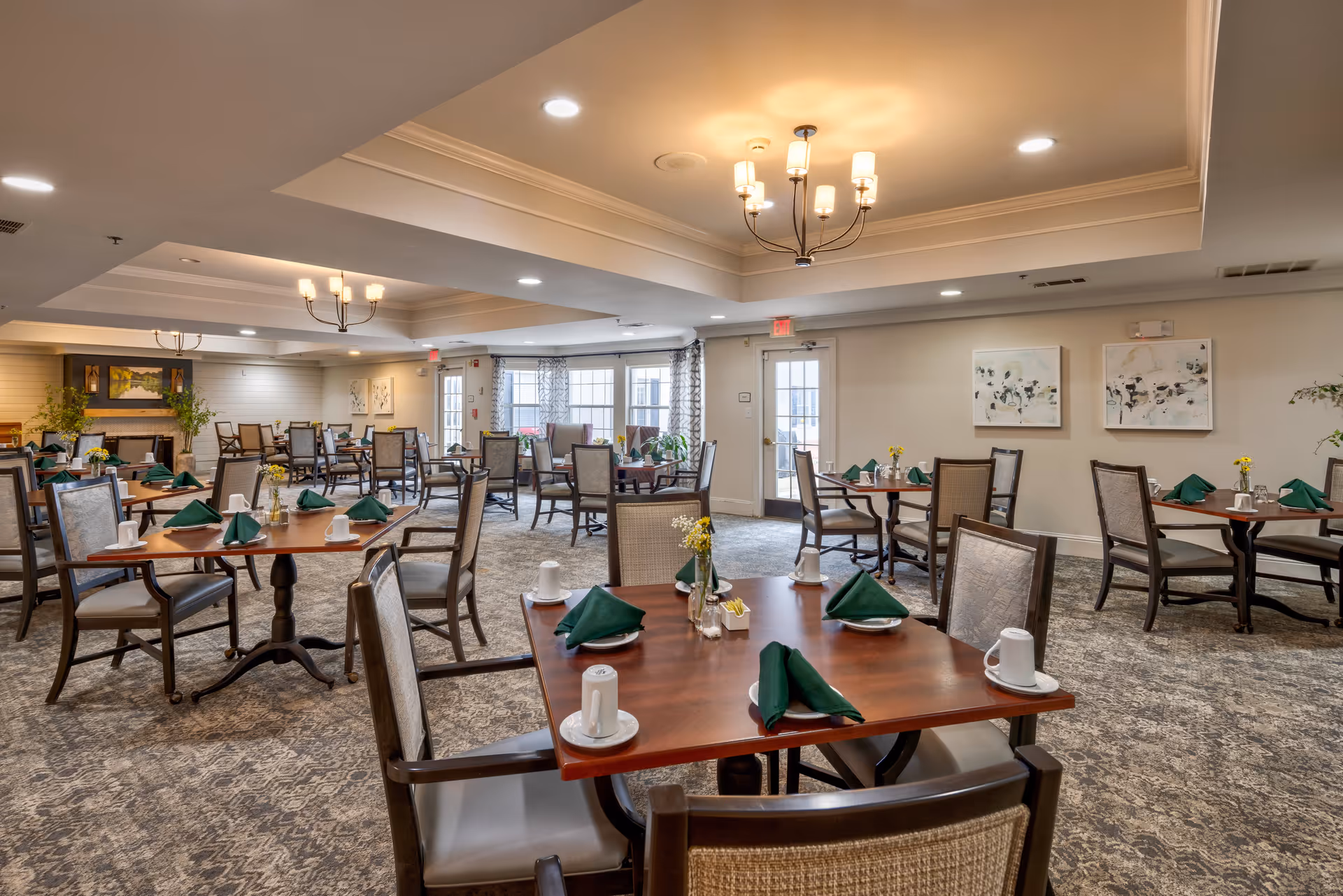 A spacious dining room with multiple wooden tables and cushioned chairs arranged neatly. Each table is set with green folded napkins, white cups, and small flower vases. The room has soft lighting from ceiling chandeliers and recessed lights, with large windows and a door letting in natural light. The walls are decorated with framed artwork and there is a fireplace in the background.