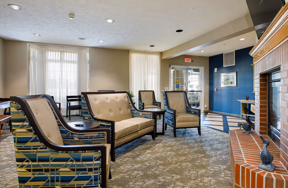 A senior living facility common area with patterned armchairs and a loveseat arranged around small tables. The room has large windows with sheer white curtains, a brick fireplace with decorative sculptures on the hearth, and a blue accent wall with a framed picture. The floor is carpeted with a patterned design, and there is an exit door in the background.