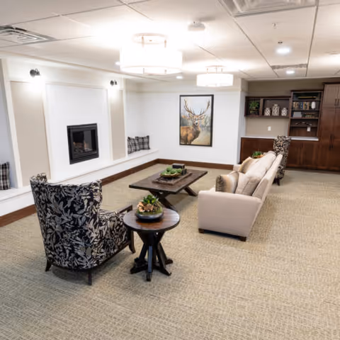 A cozy senior living facility common area with a beige sofa, two patterned armchairs, a wooden coffee table, and a side table with a small plant. The room features a built-in fireplace, a large painting of a deer on the wall, and built-in wooden shelves with decorative items. The space is well-lit with ceiling lights and has a neutral carpeted floor.