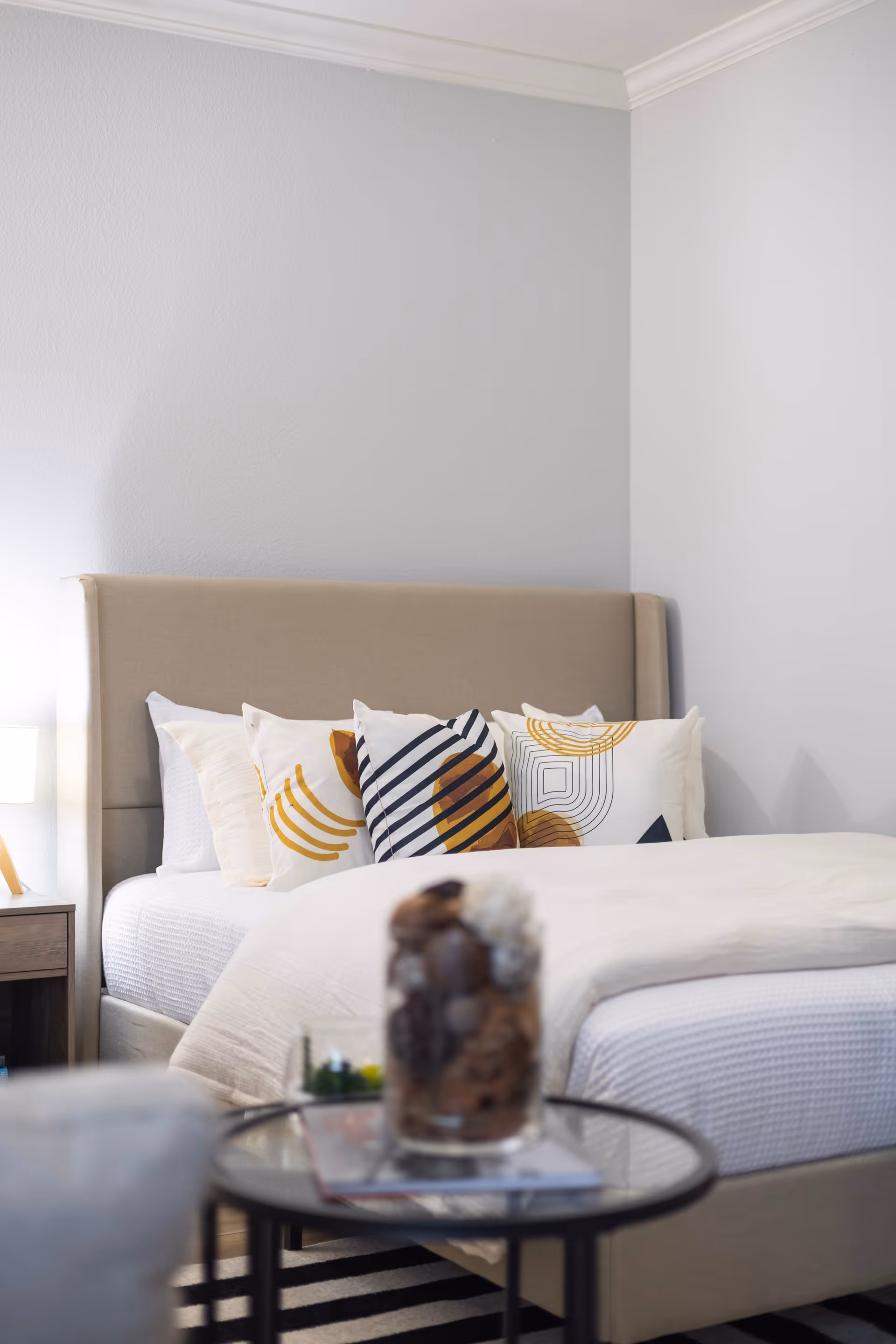 A neatly made bed with a beige upholstered headboard and white bedding, decorated with several patterned throw pillows in white, yellow, and black. A bedside table with a lamp is visible on the left side, and a round glass-top table with decorative items is in the foreground.