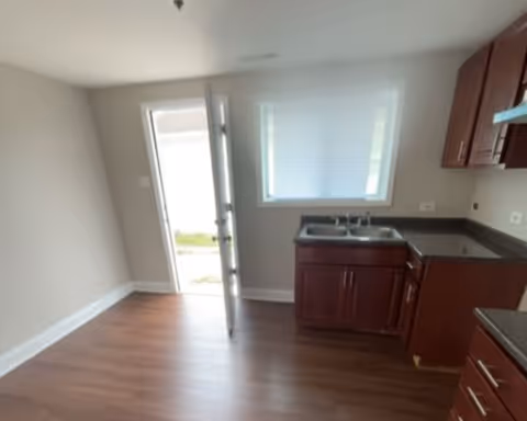 Interior view of a small kitchen area with wooden cabinets, a double sink under a window with blinds, and an open door leading outside. The floor is wood laminate and the walls are light-colored.